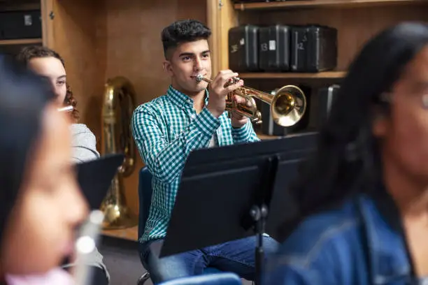 Confident male student playing trumpet in musical class. Aspiring teenage boy is learning wind instrument. He is practicing with friends in high school.