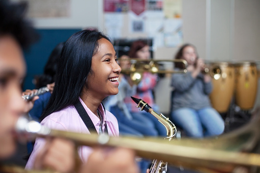 Cheerful Mixed Ethnicity Asian teenage girl sitting with saxophone while looking away. Happy female student is learning woodwind instrument in classroom. She is with friends at high school.