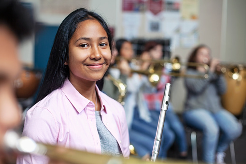 Cheerful Mixed Ethnicity Asian teenage girl sitting with flute while looking away. Happy female student is learning woodwind instrument in classroom. She is with friends at high school.