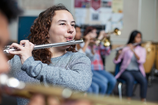 Confident female brunette student practicing flute with brass band. Teenage girl is looking away while blowing into woodwind instrument. She is playing music with friends in classroom.