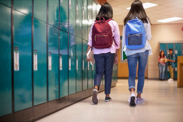 Full length of teenage girls with backpacks walking by lockers. Rear view of female friends are at illuminated corridor. High school students are at education building.