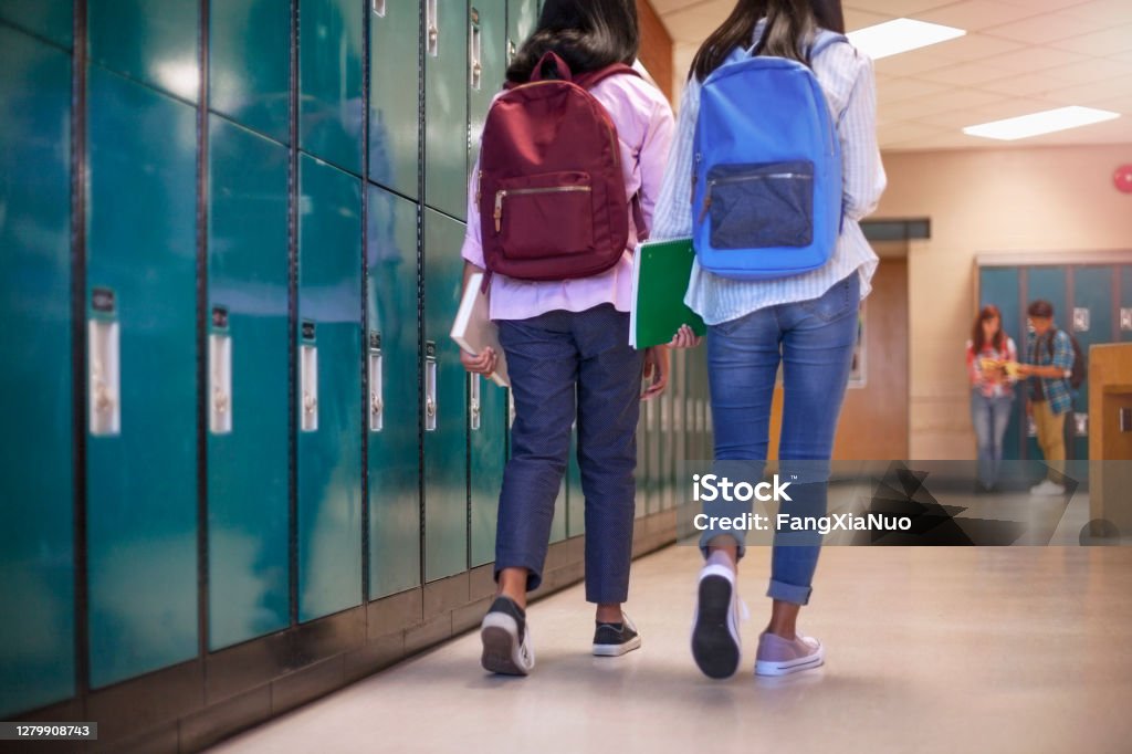 Female classmate friends with backpacks walking by lockers in school Full length of teenage girls with backpacks walking by lockers. Rear view of female friends are at illuminated corridor. High school students are at education building. School Building Stock Photo