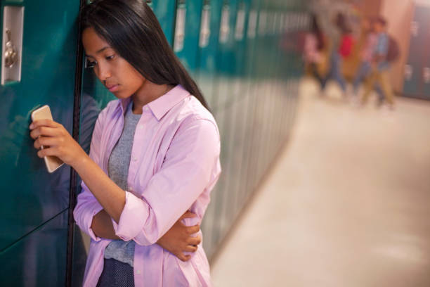 Lonely teenage Asian female student leaning on locker with smart phone in school Lonely teenage girl leaning on metallic locker. Sad female student is standing in corridor. She is at high school. cyberbullying stock pictures, royalty-free photos & images