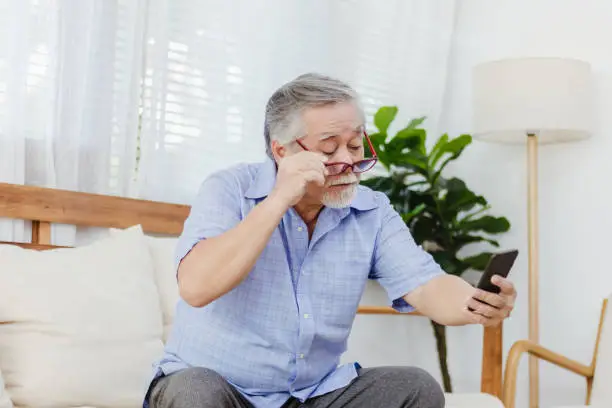 Asian senior fatigue man taking off eye glasses during using smartphone after surfing internet or social media at home. Elderly retired male eyesight problem or blurry vision from old aged
