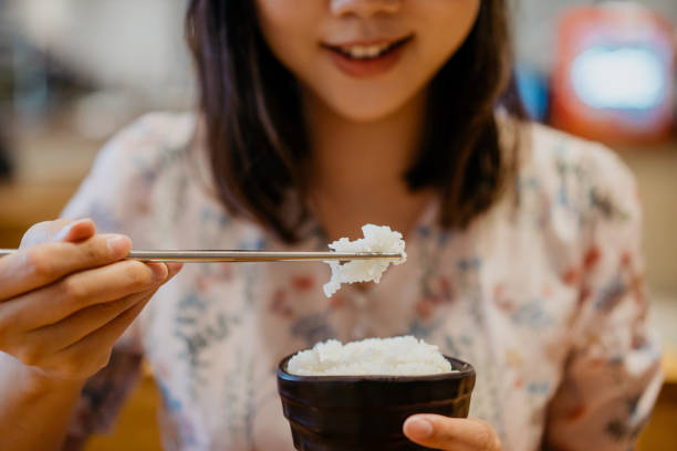 Smiling young asian woman eating a bowl of white rice Smiling young asian woman eating a bowl of white rice with chopsticks rice bowl restaurant stock pictures, royalty-free photos & images