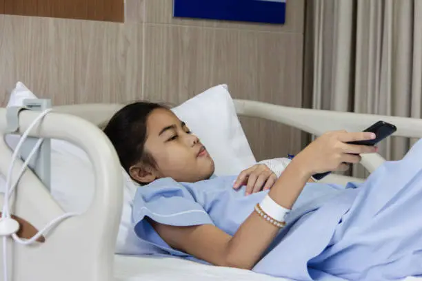 asian girl patient holding remote for select chanel at television lying on bed at hospital