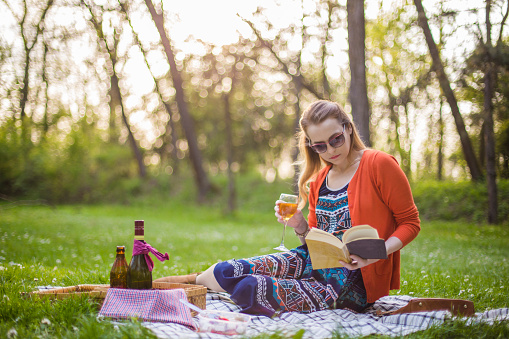 Young fashionable woman having a picnic, enjoying a sunny day, and reading a book outdoors
