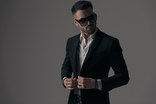 Mysterious fashion model closing his jacket, wearing suit and sunglasses while standing on gray studio background