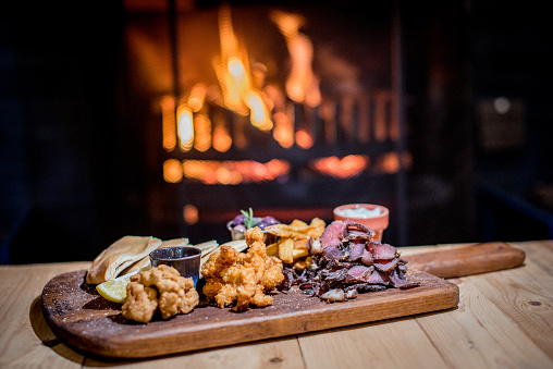 Tapas Snack Board on a winters evening with fire  in the back ground.  On the board there are some visible elements of biltong, deep friend chicken nuggets and some breads. Served on a wooden serving board.