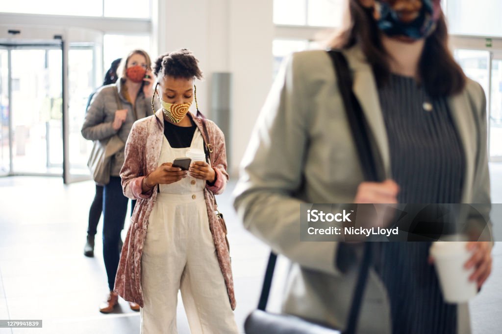 Woman in a face mask social distancing with people in a lineup Young woman in a protective face mask checking her cellphone and drinking coffee while social distancing in a line with other people Waiting In Line Stock Photo
