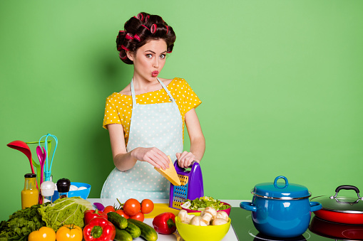 Photo of charming cute girl grate cheese table with tomato fresh, pepper ingredients make lips pouted plump wear yellow dotted t-shirt hair rollers isolated green color background