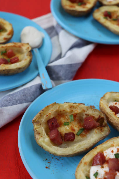 Close-up image of turquoise blue plates containing baked potato skins loaded with melted cheese, bacon cubes and sour cream, garnished with a sprinkling of chopped chives, beside blue and white stripped tea towel, red background, focus on foreground