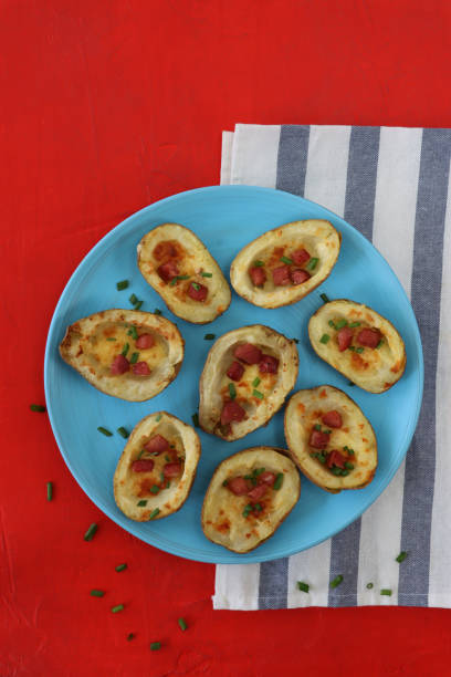 Image of group of baked potato skins loaded with melted cheese, bacon cubes and sour cream, garnished with a sprinkling of chopped chives, served on turquoise blue plate, besides blue and white stripped tea towel on red background, elevated view