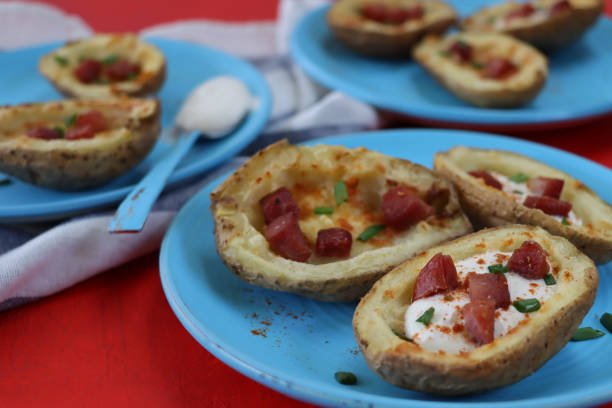 Stock photo showing close-up view of a turquoise blue plate containing a group of crispy, baked potato skins loaded with melted cheese, bacon cubes and sour cream, garnished with a sprinkling of chopped chives, sitting beside blue and white stripped tea towel on red surface. imagen de platos de color azul turquesa que contienen pieles de patata al horno cargadas de queso derretido, cubos de tocino y crema agria, adornados con un rociado de cebollinos picados, junto a la toalla de té despojado azul y blanco, fondo rojo, se cen