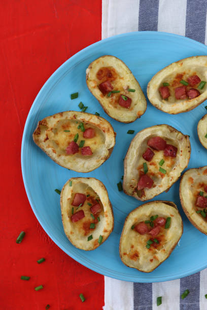 Image of group of baked potato skins loaded with melted cheese, bacon cubes and sour cream, garnished with a sprinkling of chopped chives, served on turquoise blue plate, besides blue and white stripped tea towel on red background, elevated view