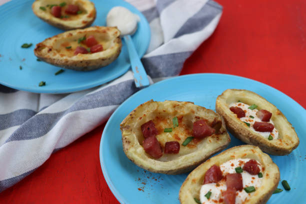 Image of turquoise blue plates containing baked potato skins loaded with melted cheese, bacon cubes and sour cream, garnished with a sprinkling of chopped chives, beside blue and white stripped tea towel, red background, focus on foreground