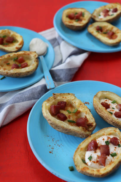 Image of turquoise blue plates containing baked potato skins loaded with melted cheese, bacon cubes and sour cream, garnished with a sprinkling of chopped chives, beside blue and white stripped tea towel, red background, focus on foreground