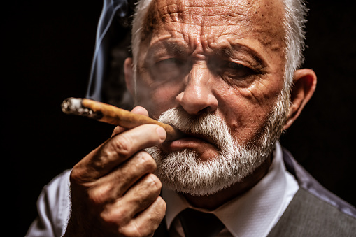 Portrait of serious senior man who is smoking cigar. Black background.
