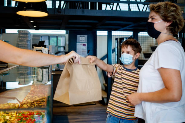 Serving customers amid Covid-19 pandemic era Male checkout server wearing protective gloves, handing take out food in paper bag, boy is reaching for bag, both customers is wearing protective masks during Covid-19 pandemic era bag lunch stock pictures, royalty-free photos & images