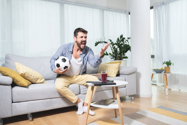 Furious football fan shouting while watching match Furious young football fan sitting on sofa holding ball and shouting while watching match football socks stock pictures, royalty-free photos & images