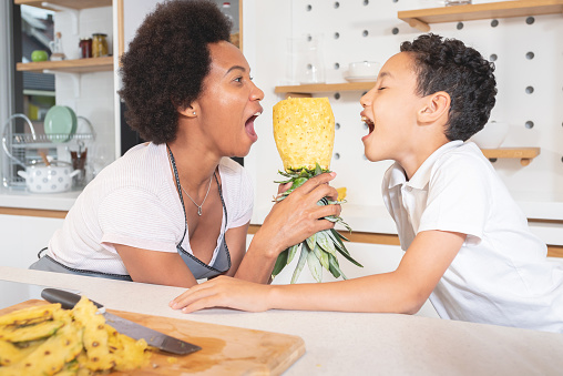 Mother and son holding peeled pineapple with mouth wide open in the kitchen