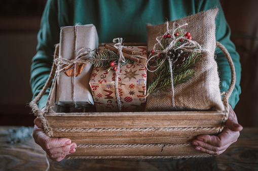 Cropped hands of unrecognizable woman puts Christmas present in wooden crate, wrapped Christmas gifts from organic recycling materials.