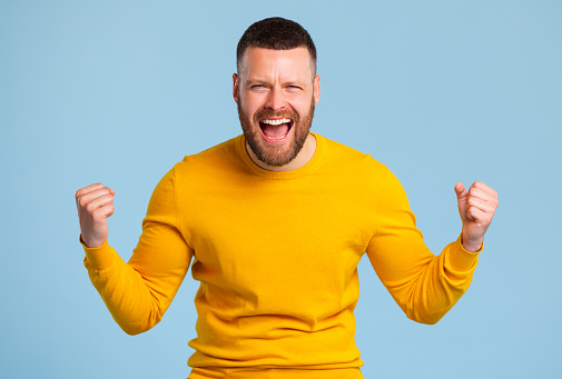happy expressive man in  yellow sweater shouts celebrating victory success and triumph on a blue background