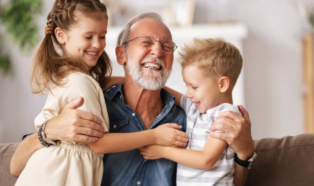 Grandfather hugging grandchildren on sofa Cheerful aged man smiling and embracing cute boy and girl while resting on couch at home together grandchild photos stock pictures, royalty-free photos & images