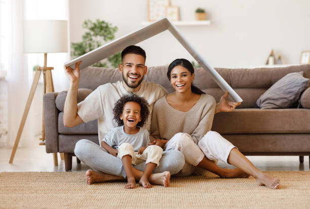 Happy family under fake roof in living room Cheerful parents with child smiling and keeping roof mockup over heads while sitting on floor in cozy living room during relocation imitation photos stock pictures, royalty-free photos & images