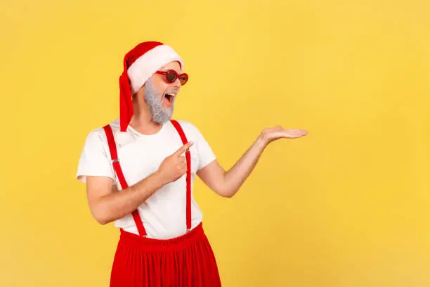 Positive elderly man in sunglasses and santa claus hat pointing finger showing freespace, advertisement area. Indoor studio shot isolated on yellow background