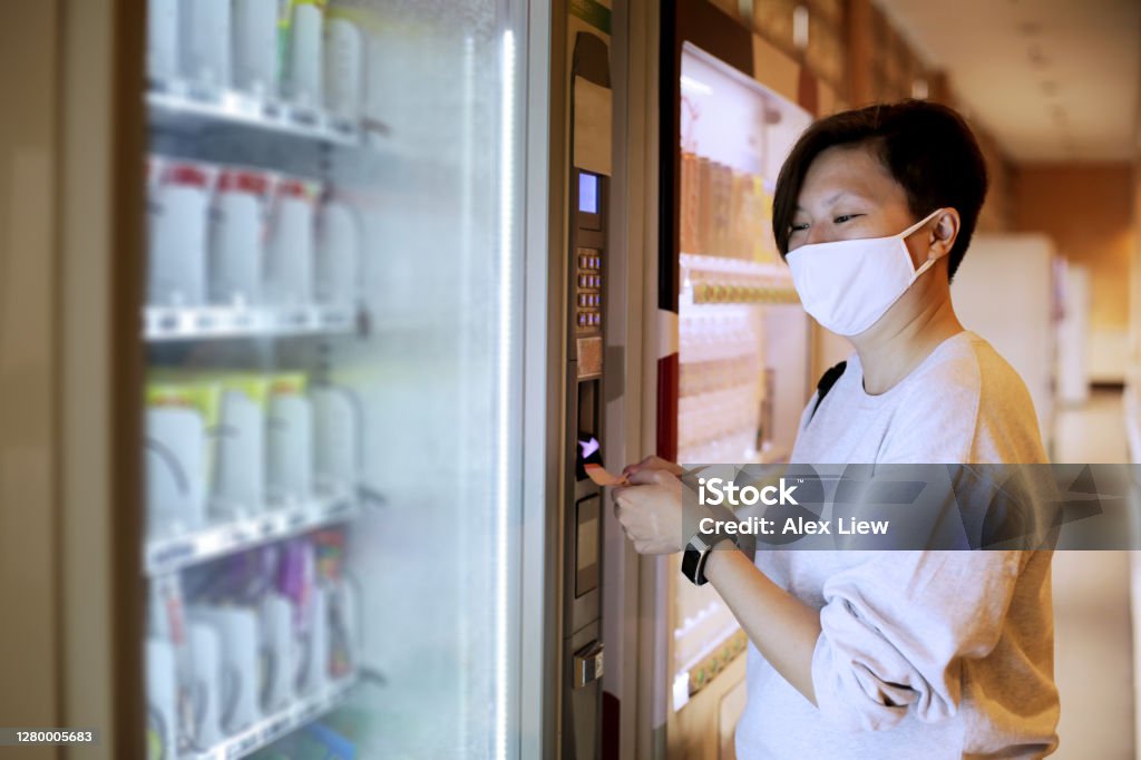 The New Norm A Chinese female adult is inserting Malaysian Ringgit (RM20 note) for payment at vending machine. She is seen wearing face mask as part of the new norm in Malaysia. Vending Machine Stock Photo