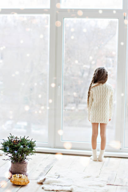 니트 흰색 스웨터 드레스를 입은 작은 행복한 소녀는 파노라마 창을 들여다보고 춤을 추며 불빛이 있는 냄비에 크리스마스 트리를 장식합니다. 새해를 기다리는 겨울 휴식. bokeh - loft apartment window apartment vehicle interior 뉴스 사진 이미지