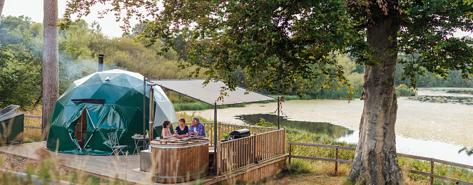 Pano shot of a family sitting together around hot tub with a lid that is being used as a table on a patio eating and drinking at a lakeside glamping site in Northumberland.