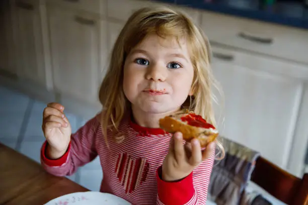 Photo of Cute funny toddler girl eats sweet bun for breakfast. Happy child eating bread roll with strawberry jam. Health food for children and kids with selfmade jelly