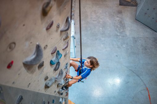 Personal perspective of determined Caucasian child sportsman suspended by rope and looking up as he scales vertical slab in modern sport climbing center.