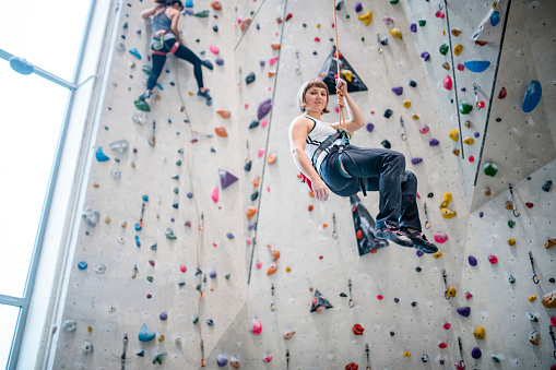 Action portrait of 23 year old Caucasian sportswoman in mid-air looking at camera as she descends from scaling climbing wall.