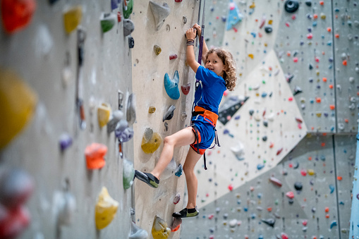 Side view of skilled 7 year old Caucasian boy gripping hand and foot holds while planning his ascent up climbing wall in modern indoor gym.