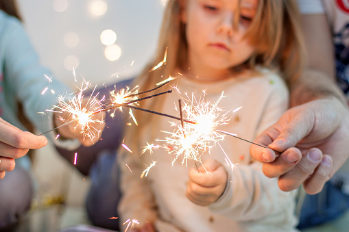 Happy family light sparklers, have fun, fool around on the background of yellow garlands of lights and bokeh Christmas tree branches. Good New Year spirit. Parents and daughters throw confetti, hug,