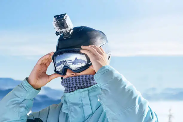 Skier with action camera on a helmet. Ski goggles  with the reflection of snowed mountains. Portrait of man at the ski resort on the background of mountains and blue sky,. Wearing ski glasses