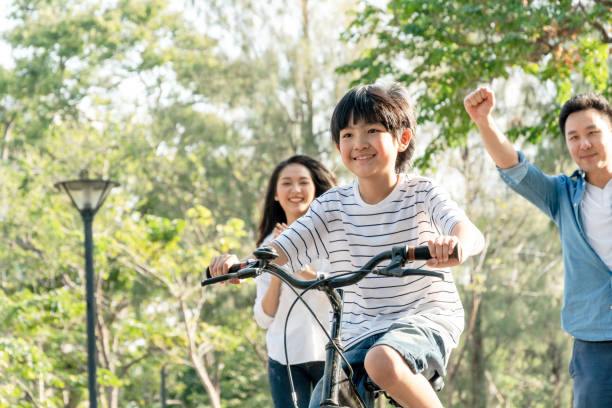 heureux père asiatique et la mère enseignent à leur fils à monter un vélo, parent joyeux lever les mains en l’air pour soutenir l’encouragement des enfants, la famille faire de l’activité ensemble au concept de parc - family bicycle cycling healthy lifestyle photos et images de collection