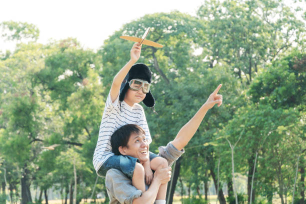 азиатский мальчик в пилотном шлеме сидит на плече отца, держа в руках самолетную игрушка. папа обучения сына и указывая руку вперед в парке.  - aspirations pilot child airplane стоковые фото и изображения