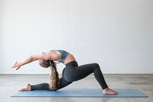 Profile view of an athletic woman doing a low lunge position yoga pose for flexibility. She is practicing in the gym set up in the garage of her home.