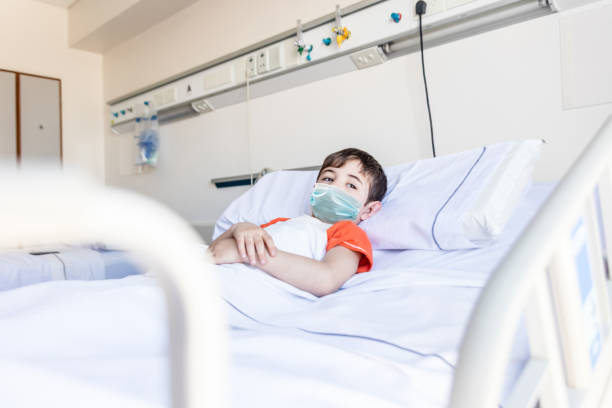 nachdenklicher kleiner junge, der auf dem krankenhausbett liegt und mit traurigkeit wegschaut und eine schützende gesichtsmaske trägt - little boys child sadness depression stock-fotos und bilder