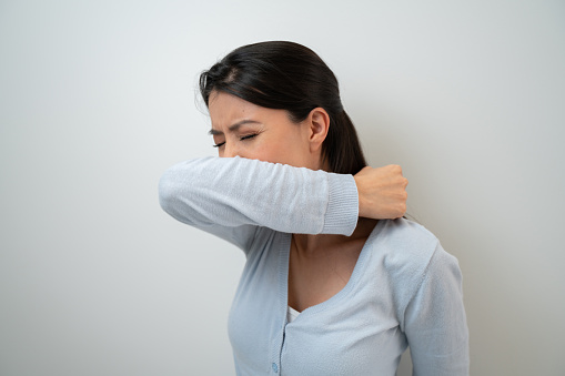 Woman wearing a facemask and sneezing in the crease of her elbow during the COVID-19 pandemic - lifestyle concepts