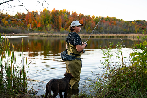 A young fly fisherman wading in a lake on a beautiful fall day.  His labrador retriever puppy is standing beside him.