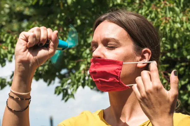 young woman cooling down with hand held ventilator while wearing a protective face mask