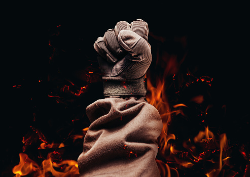 Photo of a male fist in tactical military glove and jacket on burning fire black background.