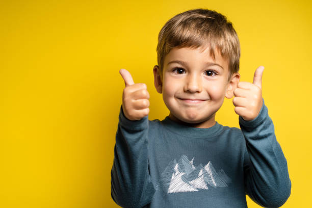 Portrait of happy small caucasian boy in front of yellow background thumbs up - Childhood growing up and achievement concept - front view waist up copy space stock photo