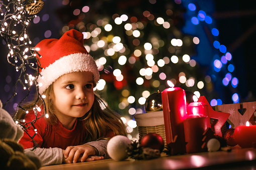 Tilt, low angle view of a cute little girl spending time on the floor of a room decorated with Christmas lights and decorations.