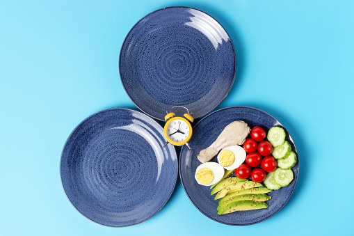 Two empty plates and a plate with food on blue background, intermittent fasting concept.